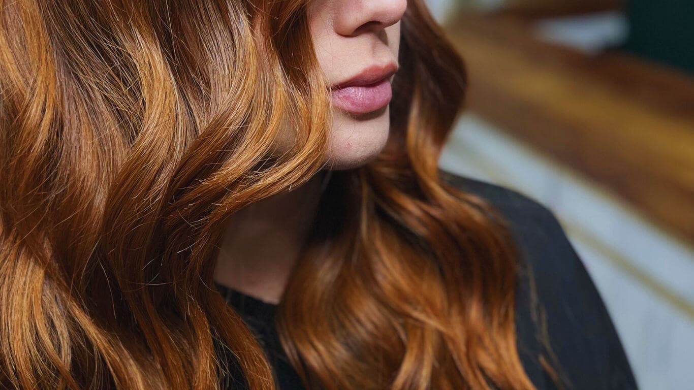 Primer plano del perfil de una mujer con el cabello largo y ondulado en tonos cobrizos, con brillo natural
